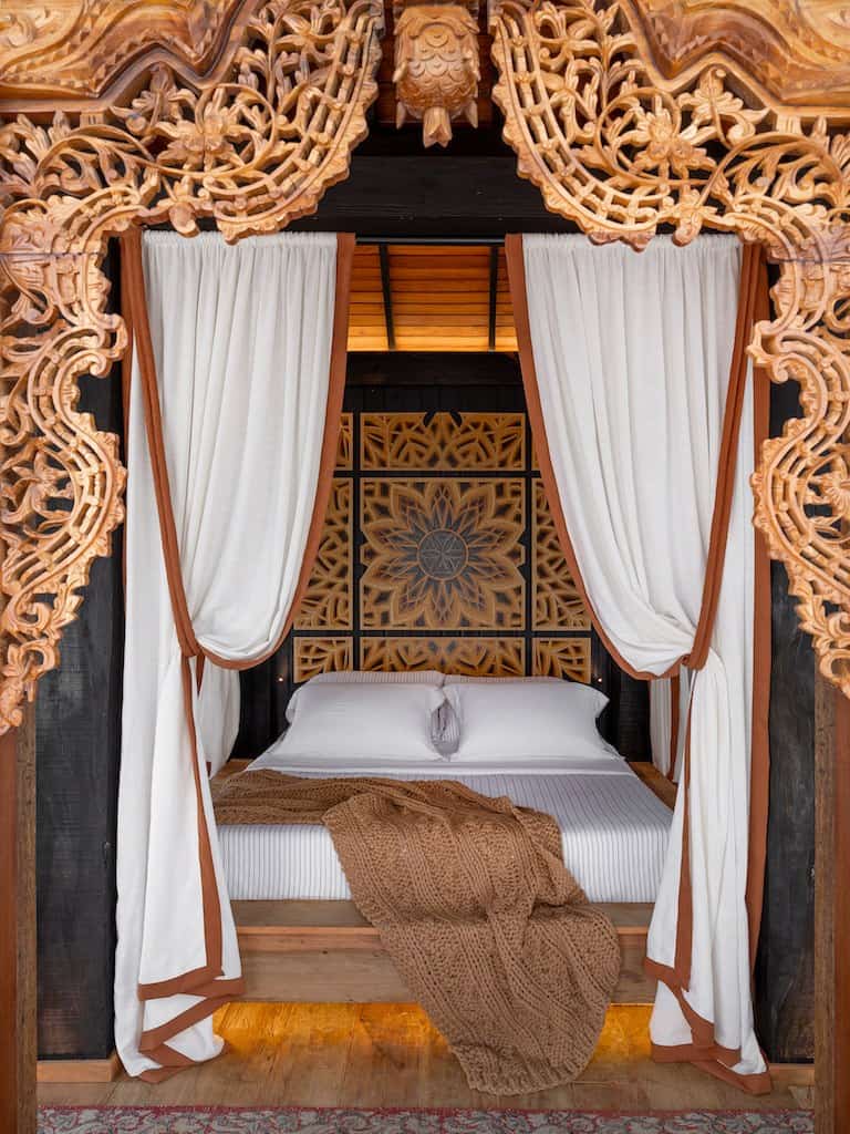 Cama emoldurada por painéis de madeira entalhada e cortinas na Reserva Ubud, de Leandro Sumar, na CASACOR SC 2026