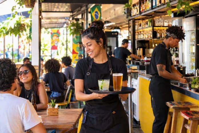 Garçonete sorridente de avental preto carrega bandeja com um drinque e um copo de cerveja em um bar movimentado, com clientes à mesa e um atendente preparando bebidas ao fundo