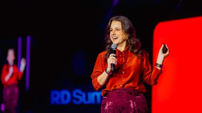 Beatriz Guarezzi, de camisa laranja, fala ao microfone em um palco com fundo escuro e painel vermelho