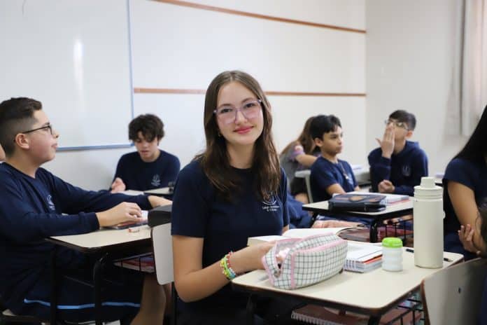 Estudante Ana Clara Bommerich, de óculos e uniforme azul-marinho, sorri sentada em sala de aula com colegas ao fundo