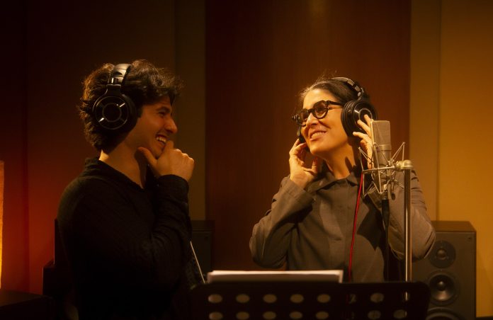 André Luiz e Mônica Salmaso sorriem de fones de ouvido diante de um microfone durante gravação em estúdio