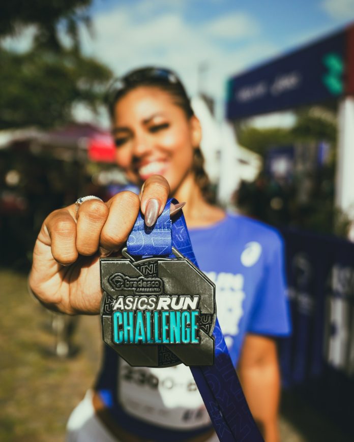 Corredora sorridente de camiseta azul segura em primeiro plano a medalha do ASICS Run Challenge apresentado pelo Bradesco, com fita azul e pórtico da prova desfocado ao fundo