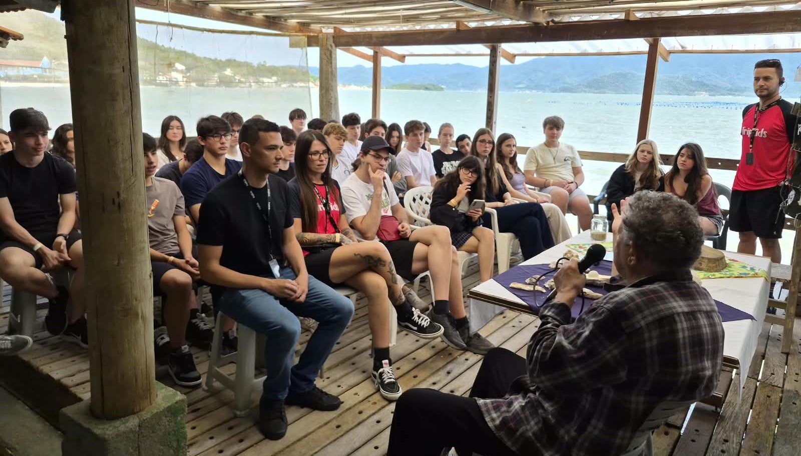 Estudantes sentados em deque à beira-mar ouvem explicação sobre maricultura no Ribeirão da Ilha