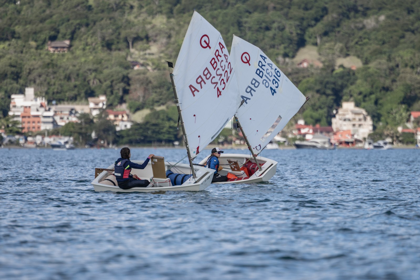 Jovens velejadores em barcos Optimist navegam lado a lado em Florianópolis