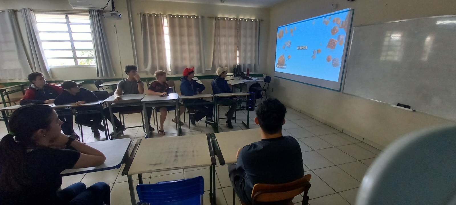 Estudantes sentados em sala de aula assistem a uma projeção durante reunião do projeto