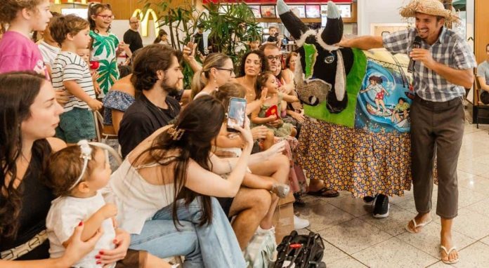 Brincante de chapéu de palha e camisa xadrez canta ao microfone ao lado do boi de pano preto e branco, diante de crianças e famílias sentadas no chão de um shopping