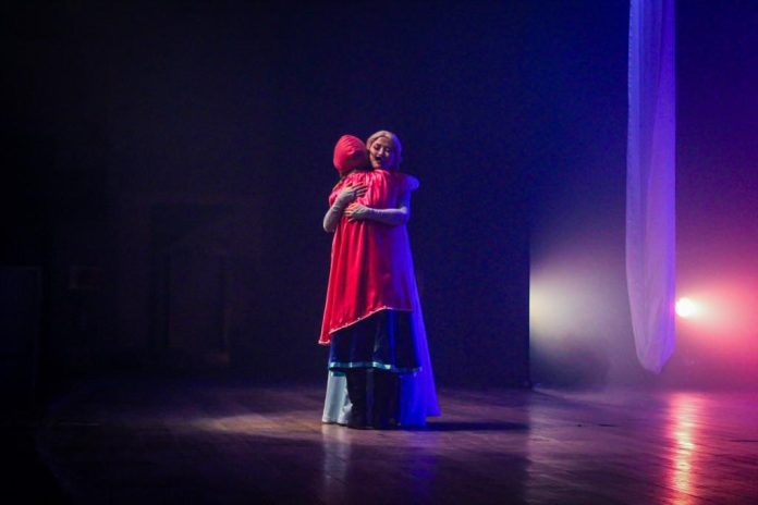 Duas personagens de um musical infantil se abraçam no centro de um palco escuro, uma com capa vermelha e capuz e outra de vestido claro, iluminadas por luz azul e rosa