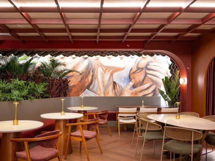 Restaurante Jardim Secreto na CASACOR SC 2026, com paredes e teto em vermelho profundo, mesas de mármore, cadeiras de madeira com estofado rosado, mural pintado de mãos e jardim tropical ao fundo