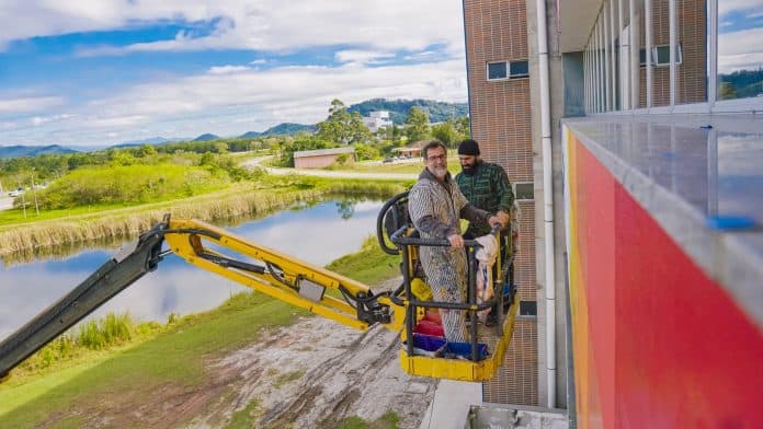 Artista Cazão e assistente em plataforma elevatória pintam faixas de cor na fachada do InPETU Hub, no Sapiens Parque