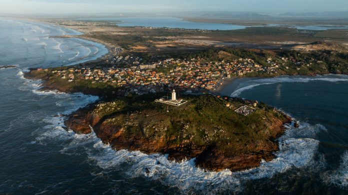 Vista aérea do litoral catarinense com farol, vila de casas e mar agitado batendo nas pedras