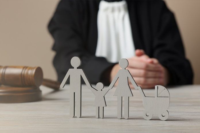 Figuras de madeira de uma família com dois adultos, uma criança e um carrinho de bebê sobre uma mesa, com um martelo de juiz e um magistrado de toga desfocados ao fundo