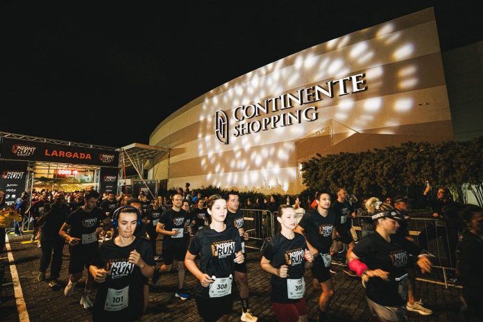 Corredores com camisetas pretas e números de peito largam à noite em frente à fachada iluminada do Continente Shopping, com o pórtico de largada à esquerda