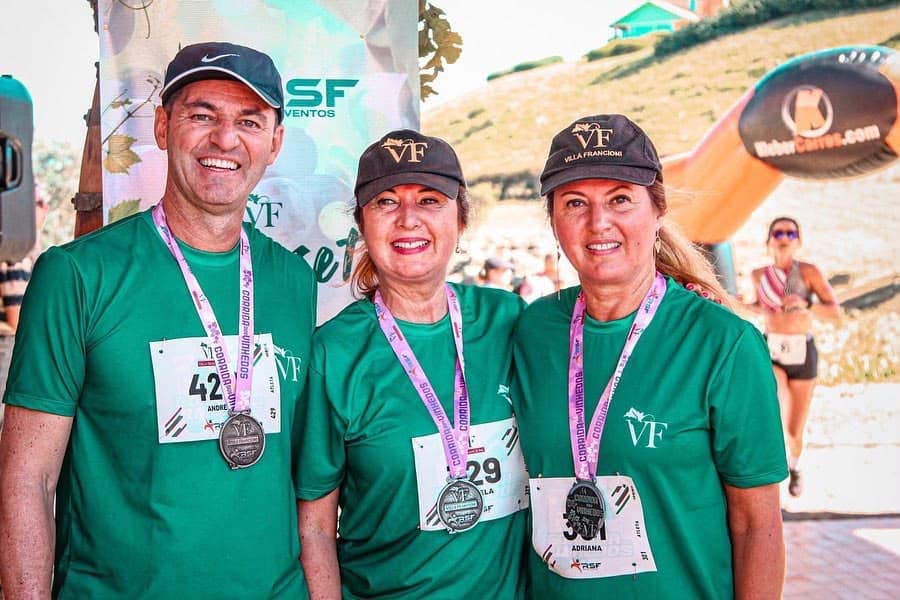 Três participantes com camisetas verdes e medalhas na Corrida dos Vinhedos