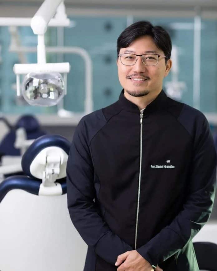 Retrato do professor e dentista Daniel Hiramatsu, de óculos e jaleco preto, sorrindo em um consultório odontológico