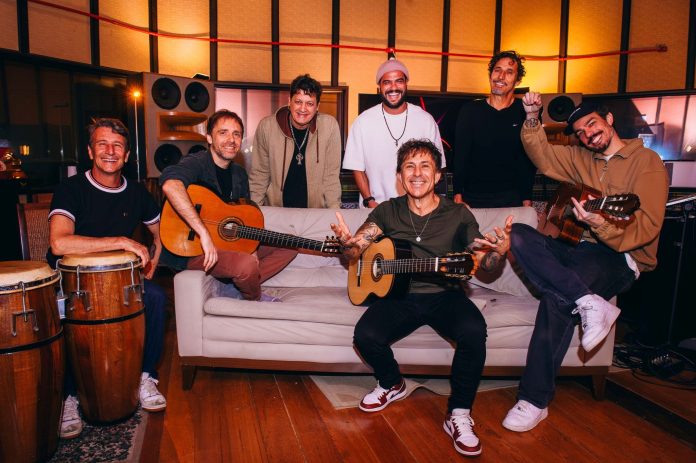Integrantes do Dazaranha e do Reis do Nada sorriem em estúdio de gravação, sentados e em pé ao redor de um sofá, com violões e congas