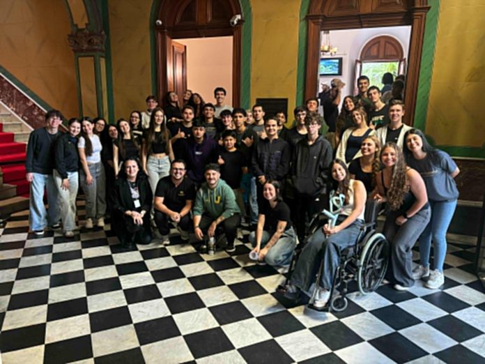 Grupo de estudantes e professores do Dazumbanho Pré-Vestibular posa no saguão de piso xadrez preto e branco do Palácio Cruz e Sousa, com uma estudante em cadeira de rodas à frente