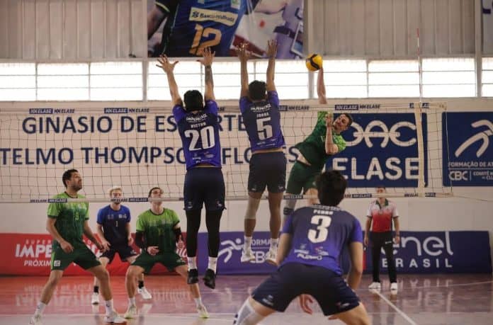 Jogadores da ELASE, de uniforme azul, saltam para bloquear o ataque de um atleta de verde da ADV Jaraguá durante a final no ginásio do clube, em Florianópolis
