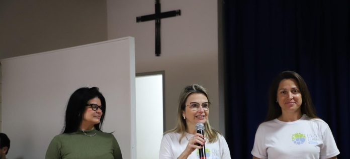 Três mulheres em palco de auditório escolar, a do centro fala ao microfone e duas vestem camisetas brancas com o logotipo Lixo Zero