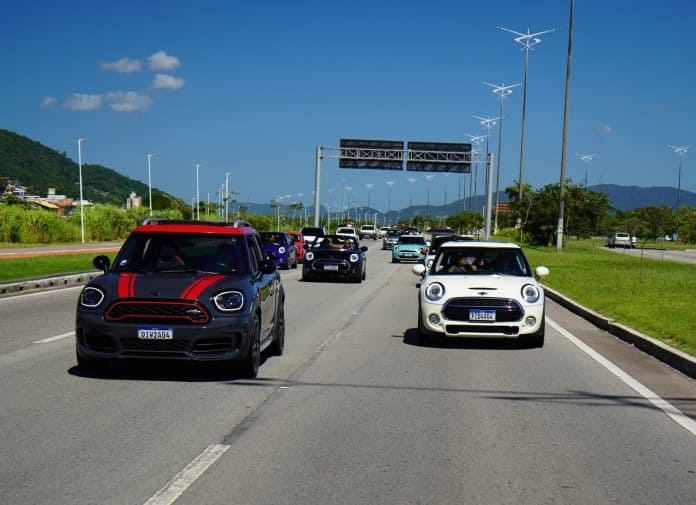 Carros Mini Cooper, um vermelho e um verde com faixas brancas, em comboio por avenida com prédios ao fundo