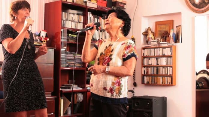 Duas mulheres cantam ao microfone em uma sala com estantes de discos, em cena da obra Karaoke – Acciones Primarias I, de Ana Gallardo