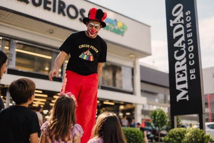 Palhaço Leleco, de perna de pau, chapéu vermelho e preto e camiseta do Circus Fever, conversa com crianças em frente ao Mercadão de Coqueiros, em Florianópolis