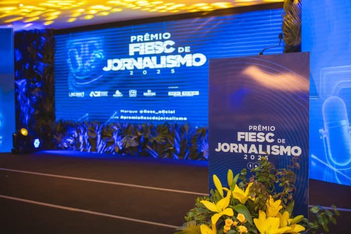 Palco do Prêmio FIESC de Jornalismo com telão azul e púlpito decorado com flores amarelas