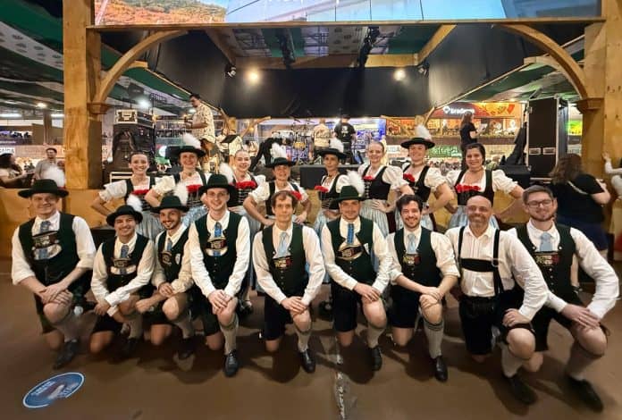Grupo folclórico em trajes típicos alemães, com chapéus e coletes, posa em corredor decorado de um centro comercial