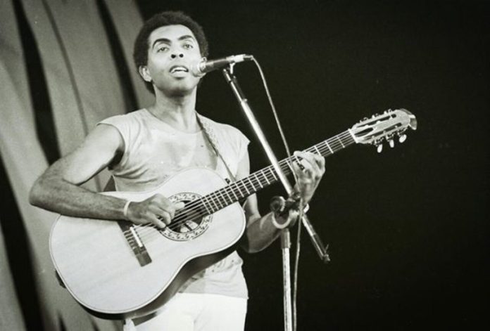 Gilberto Gil jovem toca violão e canta ao microfone em foto em preto e branco