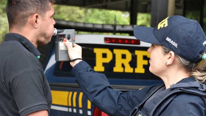 Policial rodoviária aplica teste do bafômetro em motorista ao lado de viatura da PRF