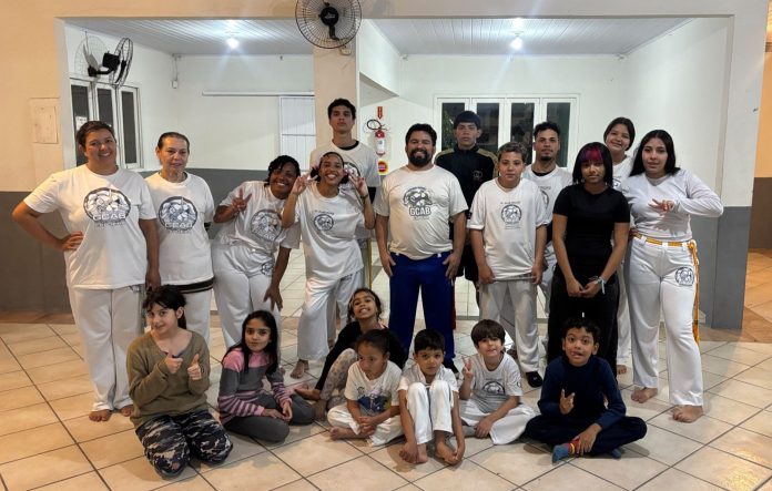 Integrantes do GCAB Capoeira, adultos e crianças, posam juntos com camisetas do grupo em sala de treino