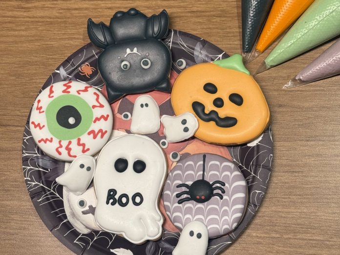 Bolachas decoradas de Halloween com abóbora, fantasma, olho e aranha em um prato, ao lado de sacos de confeitar