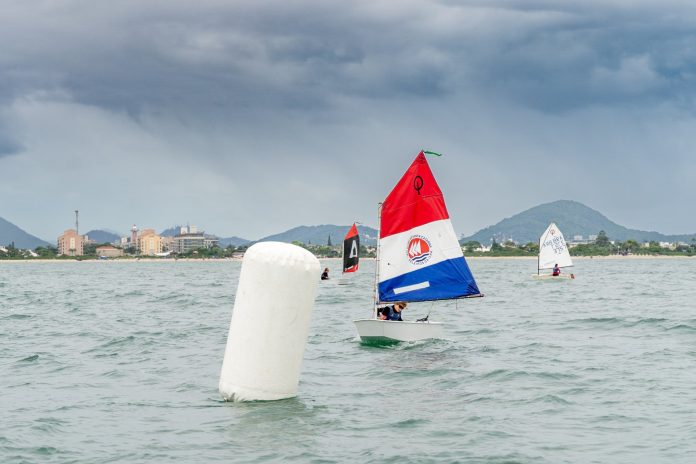Barcos da classe Optimist velejam na Baía de Jurerê, em Florianópolis, com boia em primeiro plano