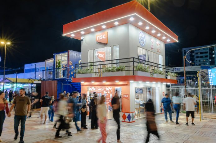 Espaço iluminado da NSC na Vila Germânica, em Blumenau, à noite, com público circulando à frente