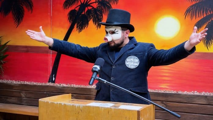 Personagem Napoleon, de cartola, máscara de porco e casaca, discursa em púlpito diante de um painel com pôr do sol e coqueiros