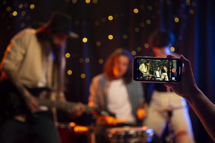Banda toca ao vivo em bar com luzes desfocadas ao fundo enquanto uma pessoa grava o show com o celular