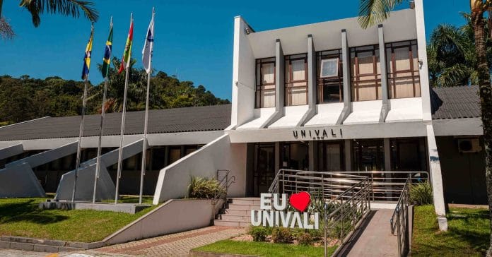 Fachada do prédio da Reitoria da Univali, com o letreiro Eu amo Univali, mastros com bandeiras e palmeiras