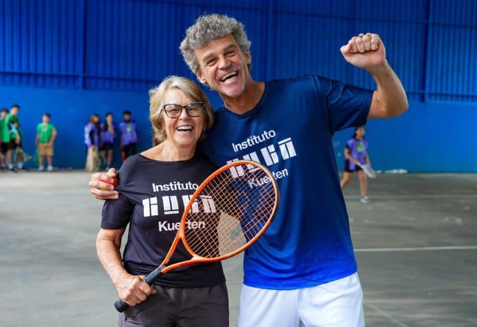 Alice Kuerten e Gustavo Kuerten sorriem e comemoram em quadra de tênis, ela segurando uma raquete laranja