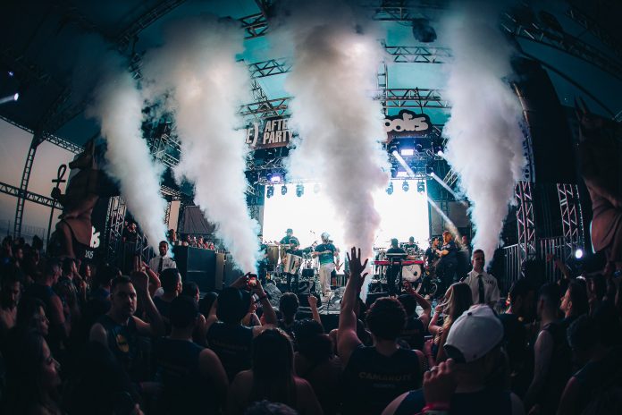 Público com as mãos para o alto diante do palco do After Camarote do Folianópolis, com jatos de fumaça branca e banda tocando sob a estrutura metálica iluminada