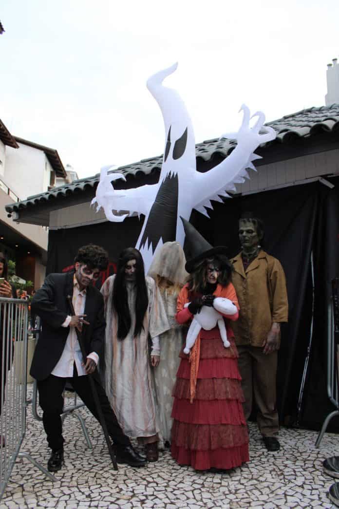 Personagens fantasiados de Halloween, com bruxa, noiva fantasma e Frankenstein, posam diante de um fantasma inflável em Jurerê Internacional