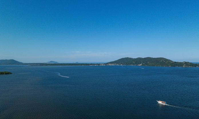 Vista aérea de baía em Florianópolis com mar azul, morros ao fundo e embarcação navegando