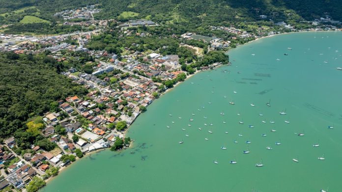 Vista aérea de bairro litorâneo em Florianópolis com casas junto ao morro e mar verde-azulado com dezenas de barcos ancorados