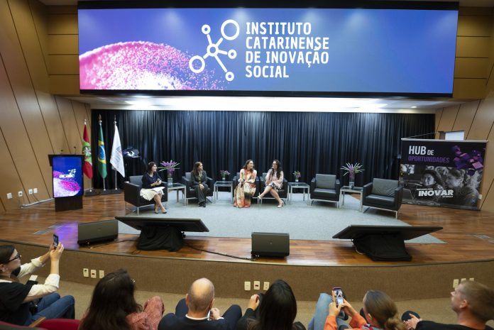 Painel com quatro mulheres sentadas em poltronas no palco de um auditório, sob telão do Instituto Catarinense de Inovação Social