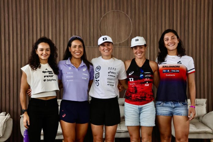 Cinco triatletas profissionais posam lado a lado, sorrindo, em frente a um painel de madeira com o logo do IRONMAN, após a coletiva em Jurerê Internacional
