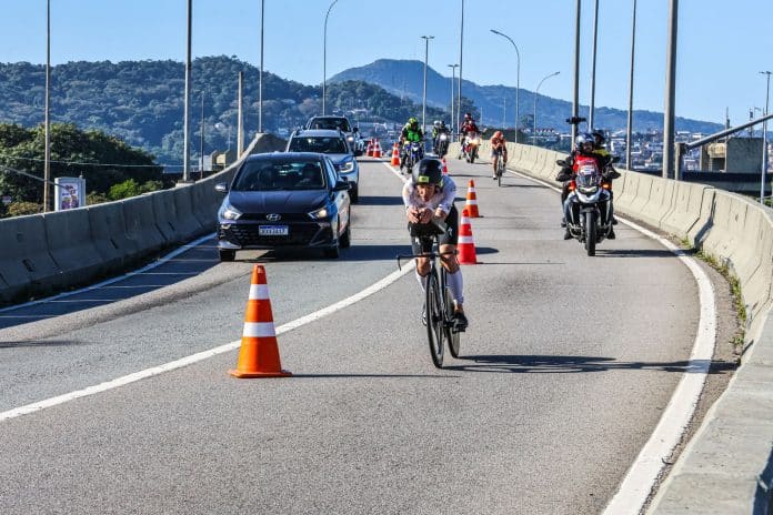 Ciclista do IRONMAN 70.3 Florianópolis pedala em faixa sinalizada com cones ao lado de carros e motos em via compartilhada