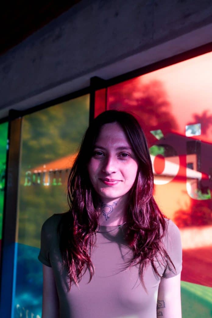 Joanna Leoni, artista e pesquisadora do Faísca – Arquivo da Memória Trans de Santa Catarina, em retrato diante de painéis de vidro coloridos