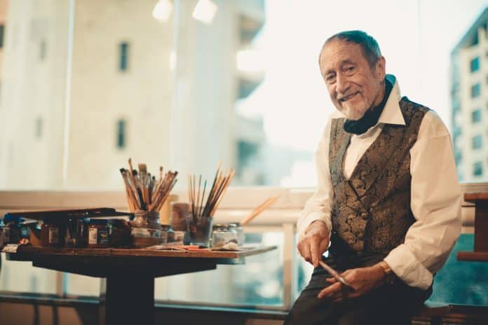 O artista Juarez Machado, de colete estampado e lenço no pescoço, sorri sentado ao lado de mesa com pincéis e tintas