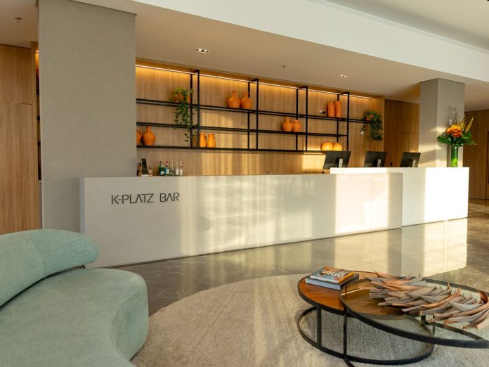 Recepção do K-Platz Hotel com balcão claro, sofá verde e mesa de centro com revistas