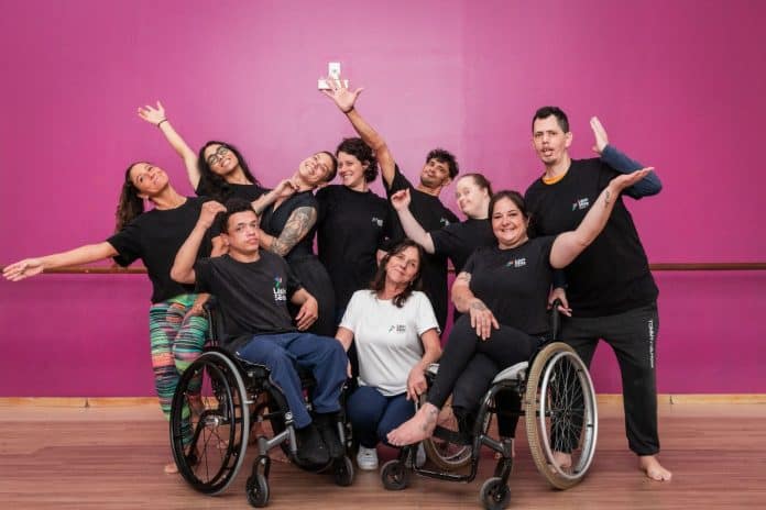 Elenco da Lápis de Seda Cia. de Dança Inclusiva, com camisetas pretas e dois bailarinos em cadeira de rodas, posa com os braços abertos diante de uma parede rosa em sala de dança