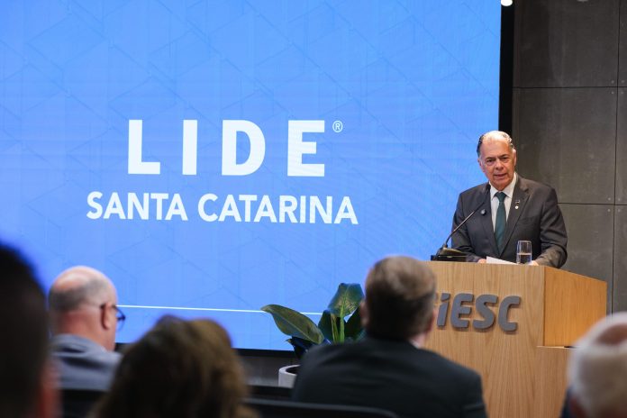 Homem de terno escuro e gravata verde discursa no púlpito de madeira com a marca FIESC, diante de um telão azul com o logotipo do LIDE Santa Catarina, durante o Encontro LIDE SC