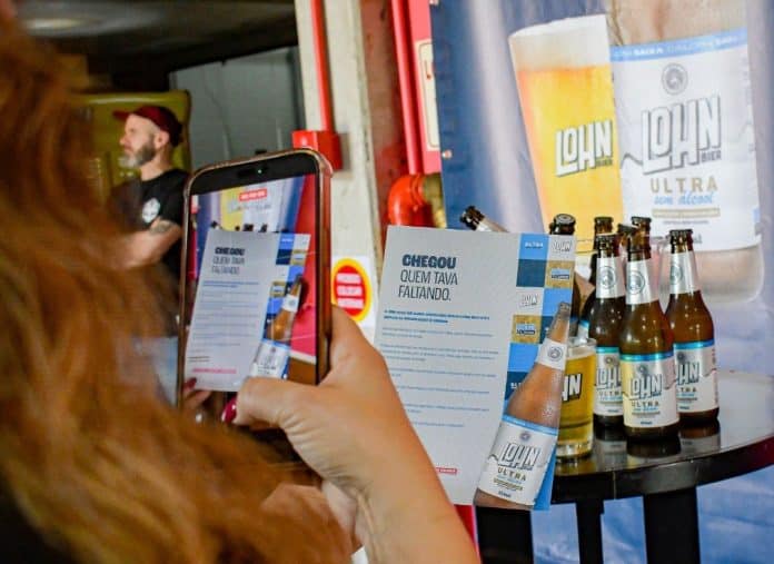 Mulher fotografa com o celular um folheto da ULTRA SEM ÁLCOOL da Lohn Bier, ao lado de garrafas da cerveja sobre uma mesa e de um painel da marca